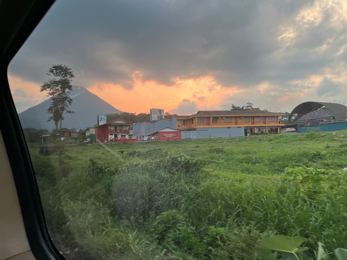 A picturesque sunset over Arenal Volcano in Costa Rica, with local buildings and lush greenery in the foreground, highlighting a travel itinerary experience.