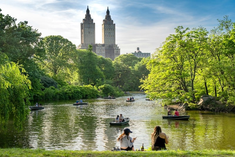 A picturesque scene in Central Park, New York City, with people enjoying a sunny day by the lake. Rowboats glide across the water, surrounded by lush greenery. In the background, the iconic twin towers of The San Remo building rise above the trees under a clear blue sky.