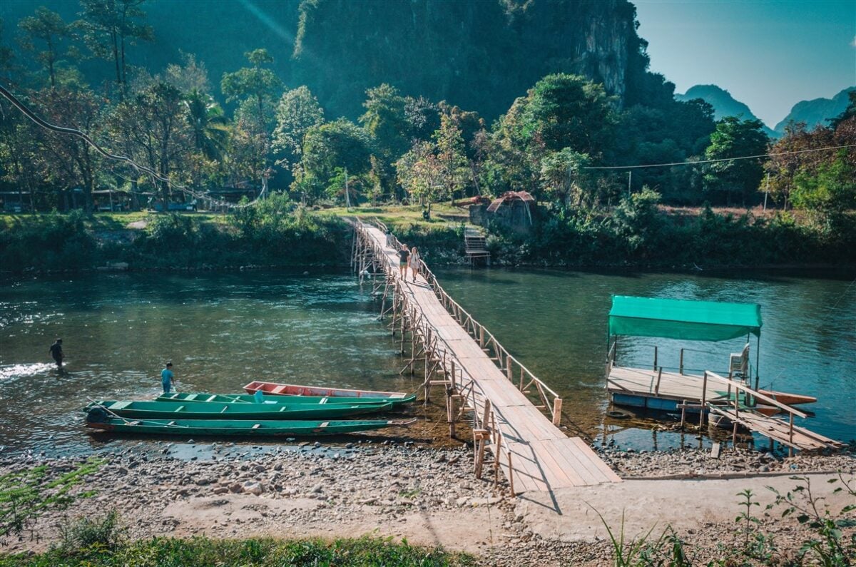 Wooden bridge over the river in Vang Vieng, Laos, with lush green trees and mountains in the background. Small boats are docked near the shore.