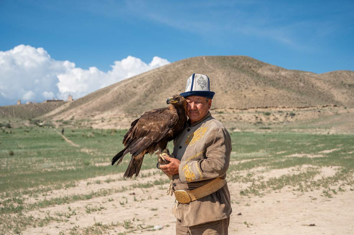 A Kyrgyz eagle hunter stands proudly in traditional attire, holding a majestic golden eagle on his arm against the backdrop of a grassy, rolling landscape and a clear blue sky. The hunter wears an ornate coat and a traditional hat, embodying the cultural heritage of eagle hunting, a revered tradition in Kyrgyzstan. The eagle, with its sharp gaze and powerful presence, symbolizes strength and the deep bond between the hunter and his bird.