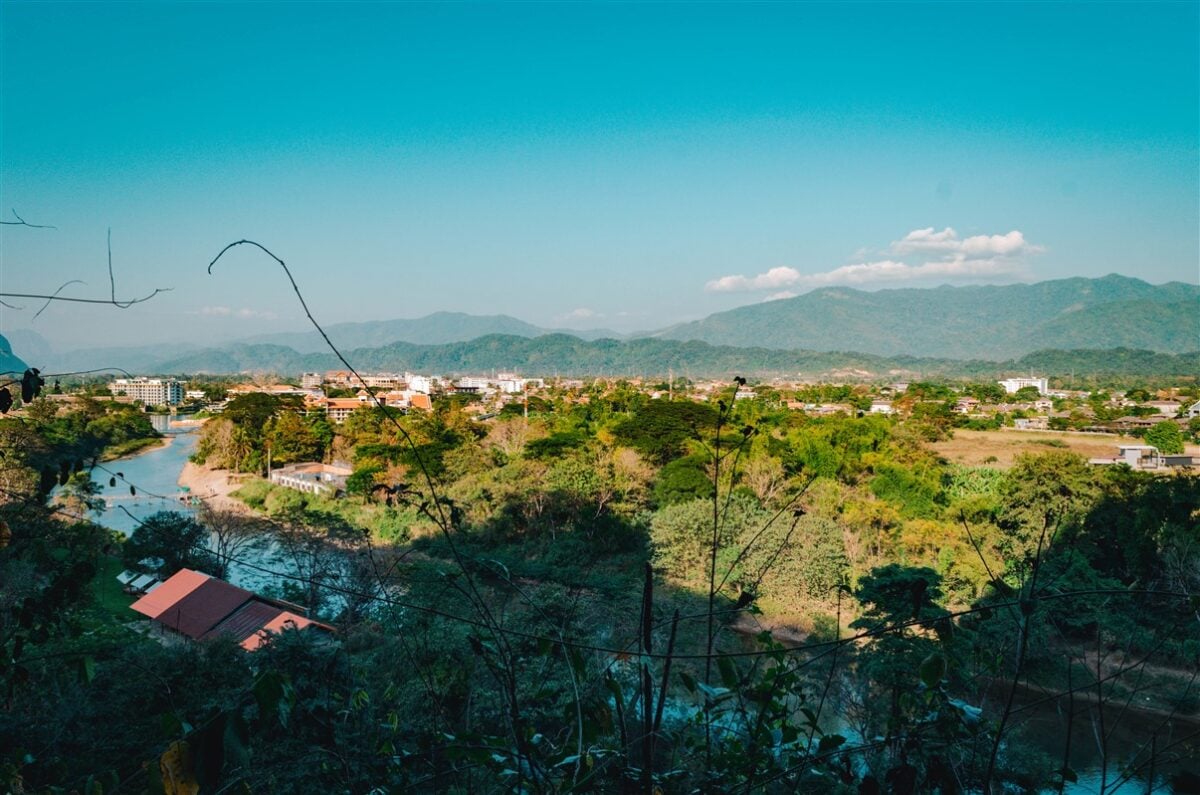 Panoramic view over Vang Vieng, Laos, from Tham Chang Cave, capturing the lush greenery, winding river, and distant mountains under a clear blue sky.