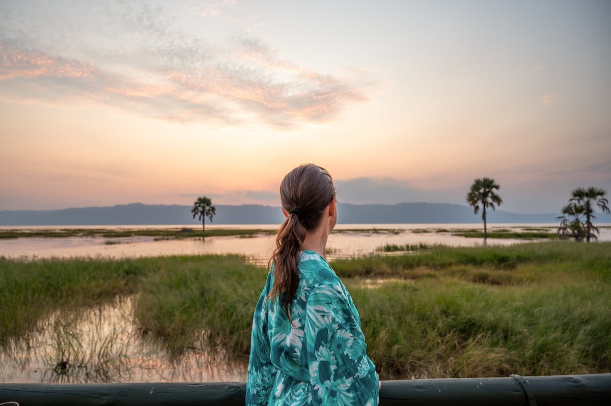 woman gazing out at a sunset over Lake Manyara surrounded by palm trees from Maramboi Tented Lodge