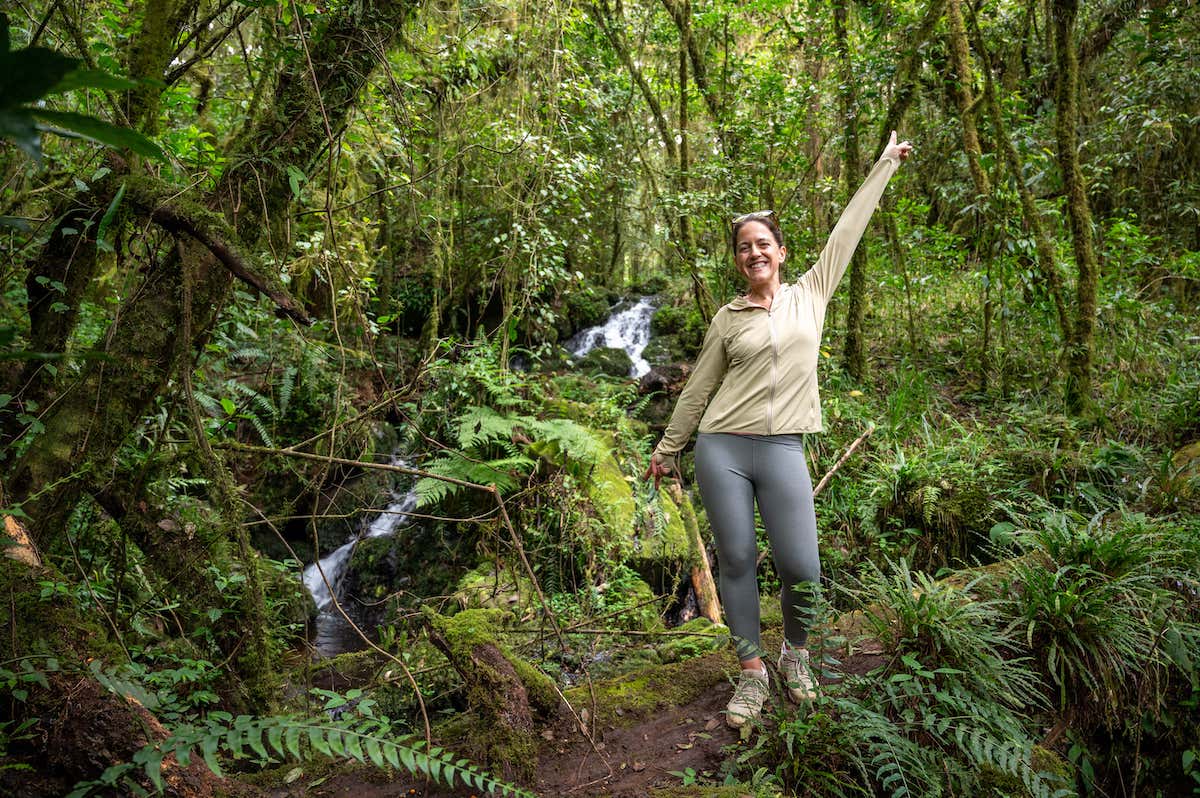 female hiker standing in front of a waterfall in Kilimanjaro National Park while hiking to Mandara Hut from Marangu Gate