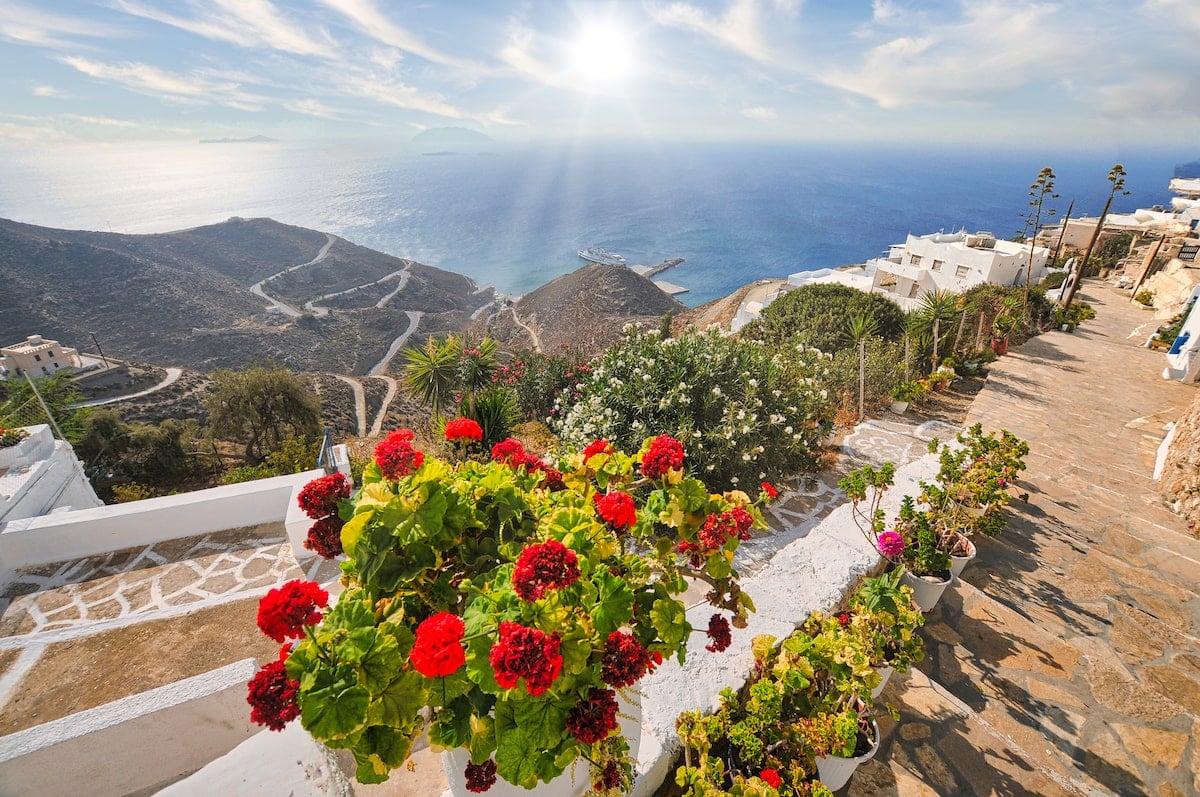 Beautiful views over Anafi Island, Greece, with a stone-paved path lined with vibrant red geraniums and other colorful plants in the foreground. The path leads to stunning panoramic views of the Aegean Sea, where sunlight shimmers over the water. The island’s rugged landscape with winding paths and white-washed buildings adds to the serene and picturesque scene, ideal for solo travelers seeking tranquility.