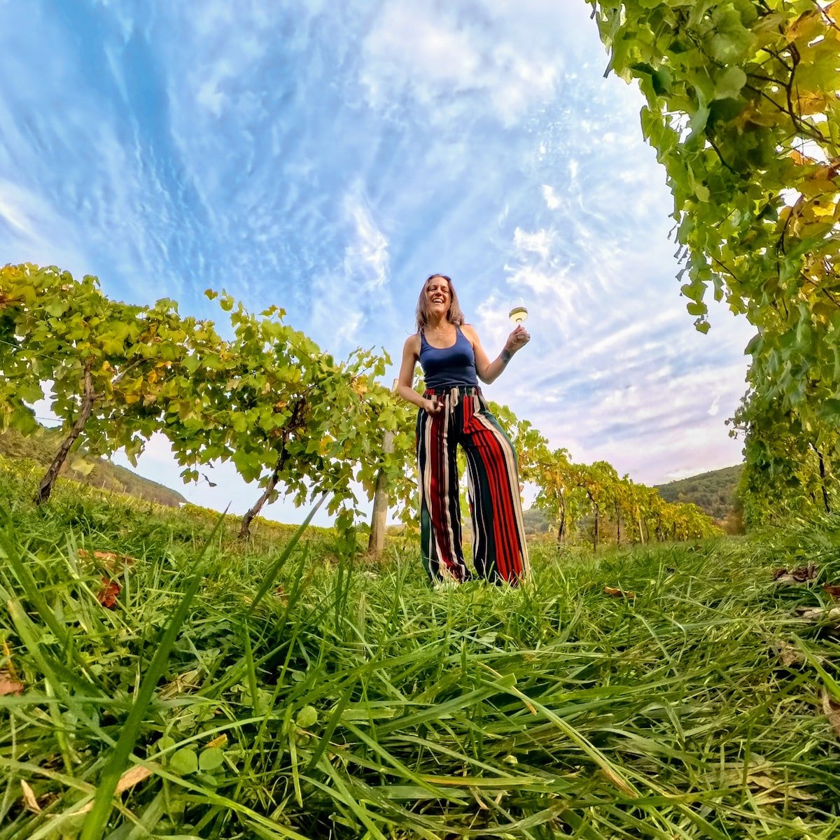 Smiling woman holding a glass of white wine, standing between rows of grapevines at Benjamin Bridge Vineyard & Winery in Wolfville, Nova Scotia, with a bright blue sky and scattered clouds overhead.