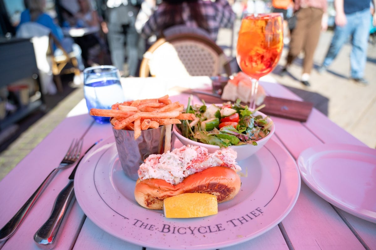 Close-up of a lobster roll served with fries, a fresh salad, and a refreshing spritz on an outdoor table at The Bicycle Thief restaurant in Halifax.