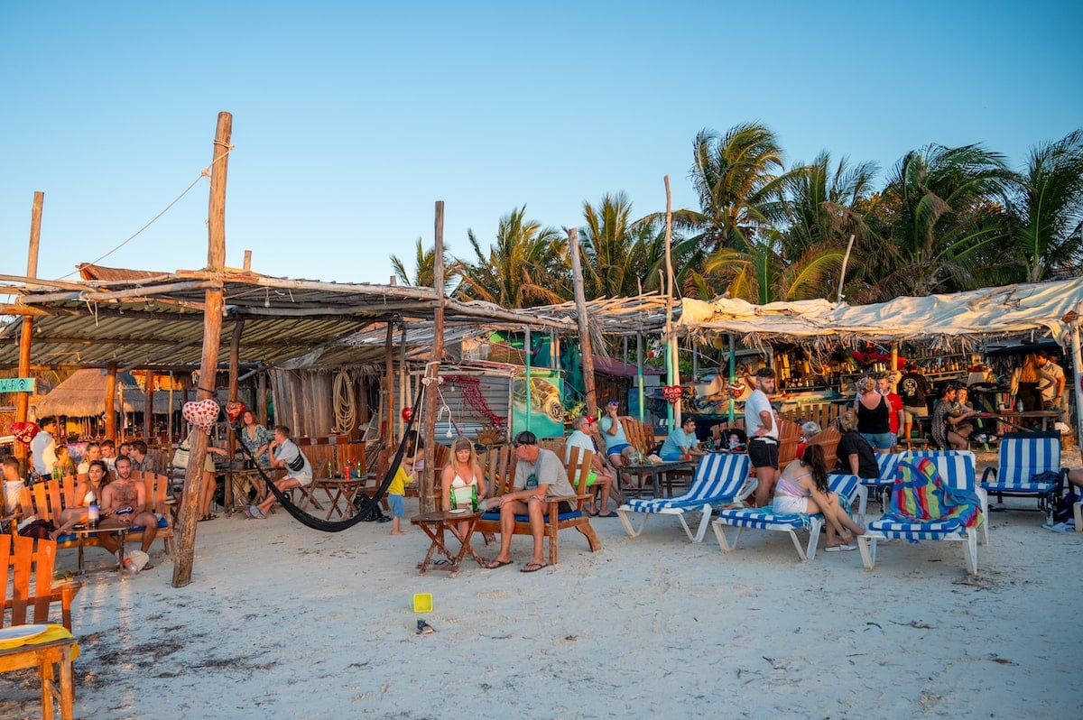 Capitán Capitán Beach Club on Holbox Island, featuring a relaxed beachside setup with wooden tables, chairs, hammocks, and blue-and-white striped lounge chairs, with guests enjoying food and drinks under rustic canopies.