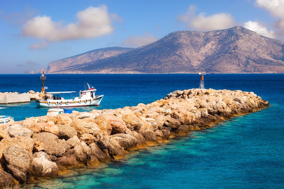 Chora fishing port in Koufonisia, Greece, showcasing a small traditional boat docked by a rocky jetty extending into the calm, clear blue waters of the Aegean Sea. In the background, a rugged island with steep slopes rises against a backdrop of a few scattered clouds, highlighting Koufonisia's peaceful and scenic charm.