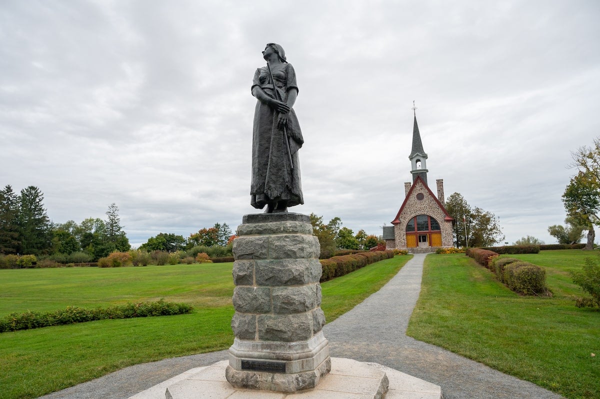 Statue of Evangeline, a character from Longfellow’s poem 'Evangeline: A Tale of Acadie,' standing on a stone pedestal with the Memorial Church in the background at Grand-Pré National Historic Site, surrounded by manicured lawns and garden pathways.