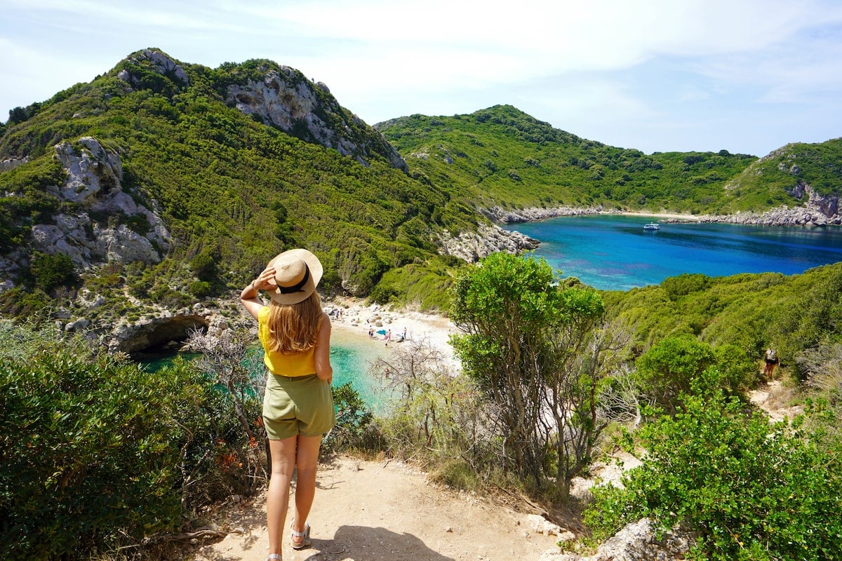 Solo female traveler in a hat and yellow top overlooking a secluded, turquoise cove surrounded by lush greenery on Corfu Island, Greece.