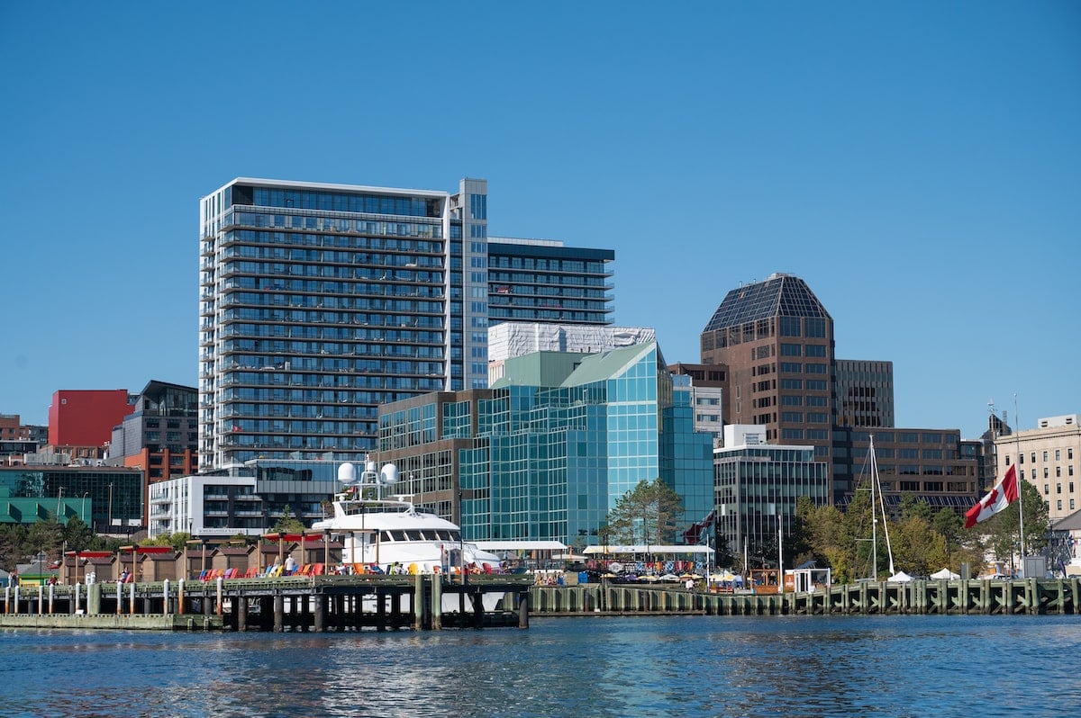 View of the modern Halifax Waterfront skyline from the Harbour Hopper tour, featuring glass buildings, high-rise apartments, and a dock lined with colorful chairs and a docked yacht under a clear blue sky.