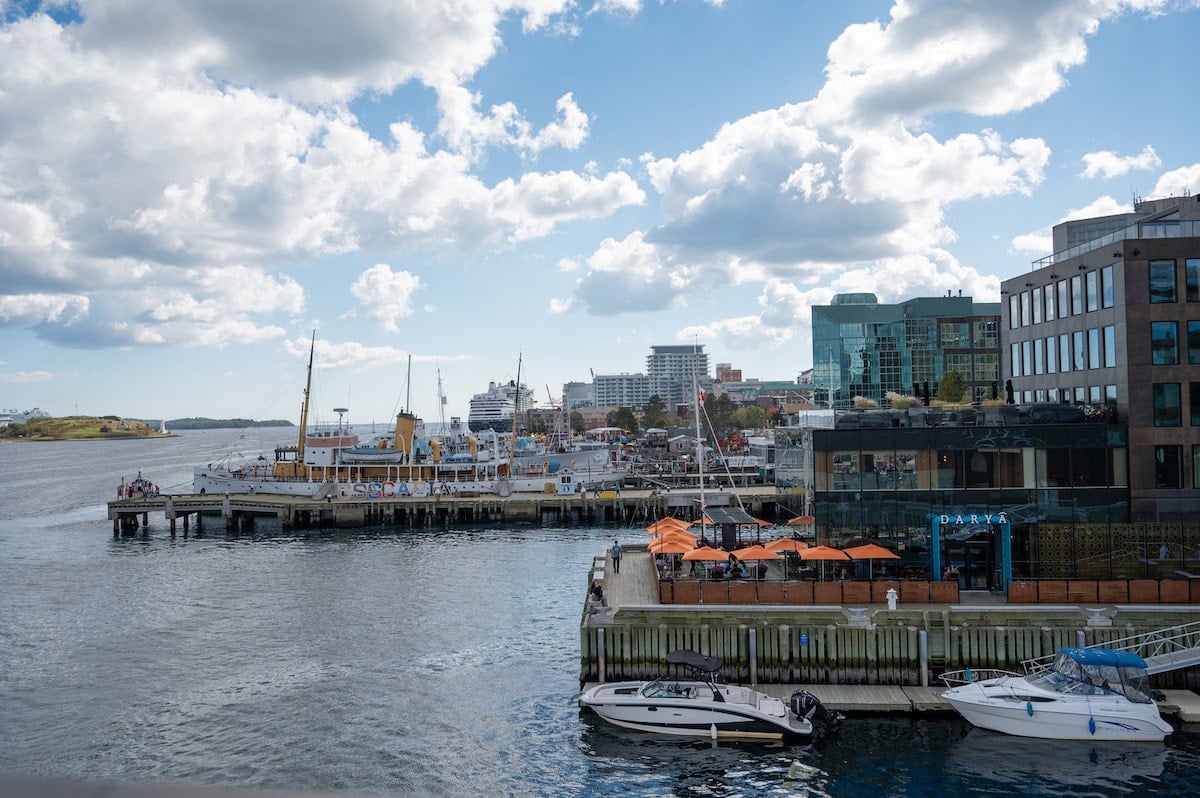 View of the Halifax Waterfront from the Queen's Marque District, showcasing the marina, docked boats, waterfront dining areas with bright orange umbrellas, and the scenic harbor under a partly cloudy sky.