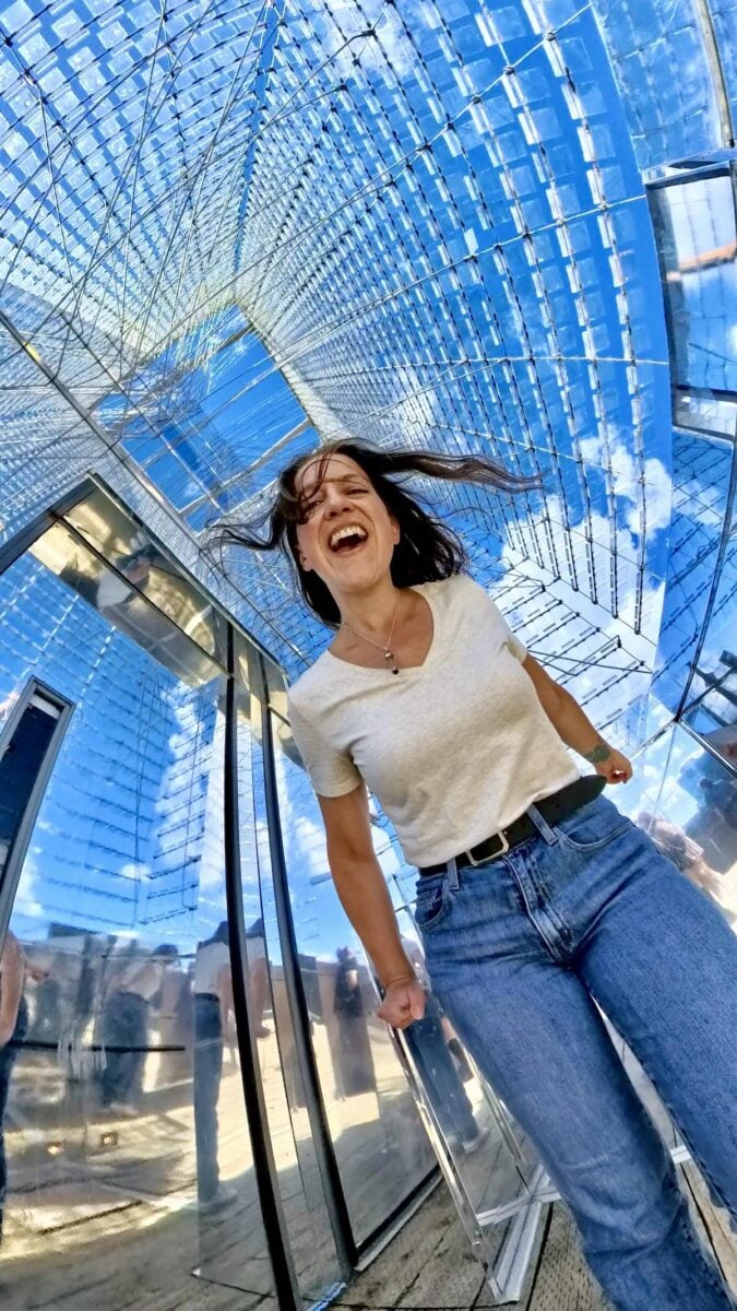 Joyful woman standing inside the Tidal Beacon installation by Ned Kahn, surrounded by reflective panels that mirror the sky and clouds, creating an immersive, dynamic visual experience.