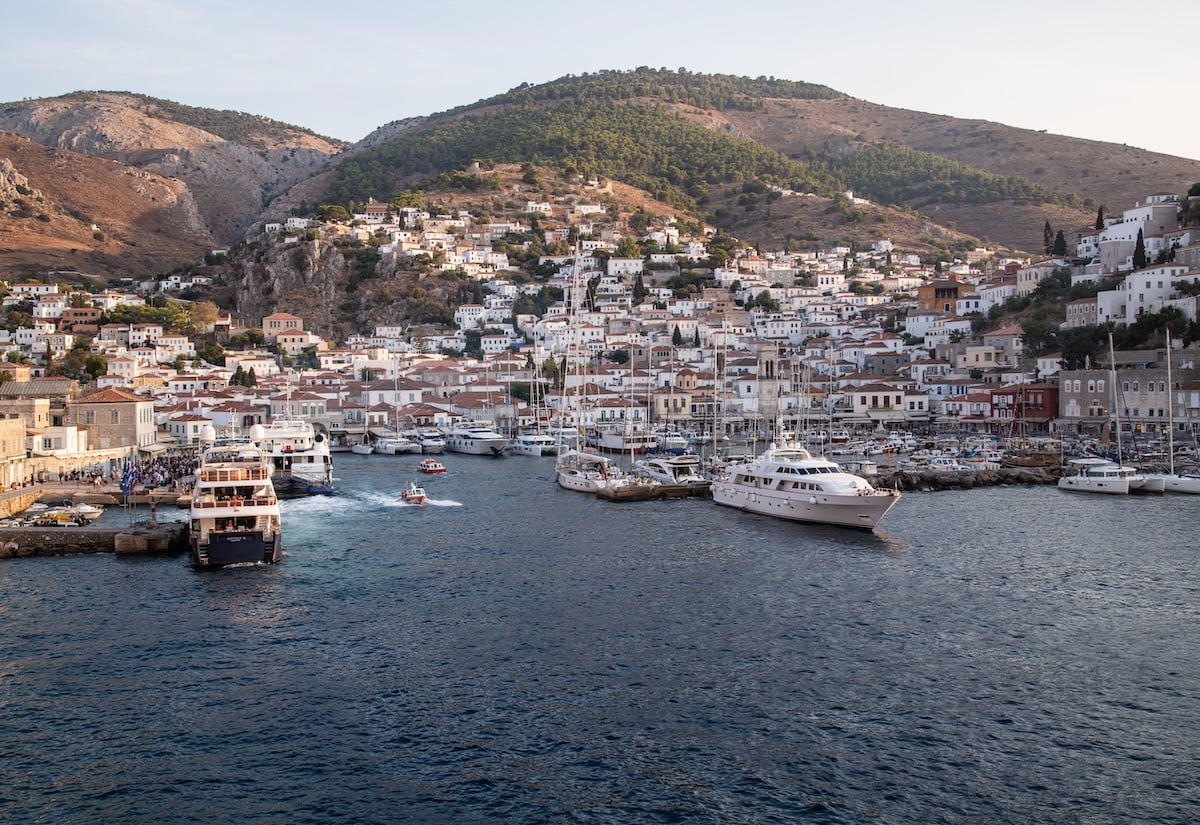 The port and town of Hydra in the Greece islands, with a bustling harbor filled with yachts and smaller boats. White-washed buildings with red-tiled roofs are nestled along the hillside, climbing up toward a backdrop of rugged mountains with patches of greenery.