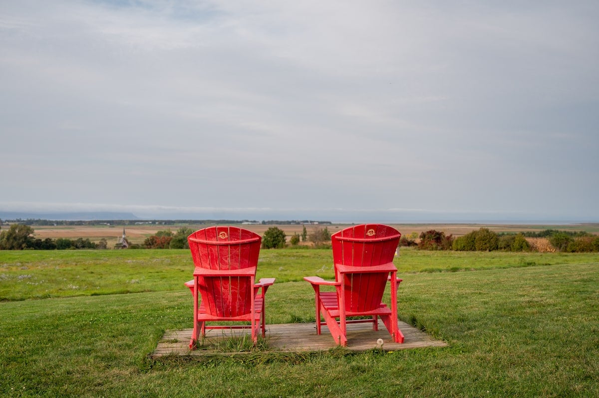 Two red Adirondack chairs overlooking the vast green fields and distant farmlands at Grand Pré UNESCO World Heritage Site, with a view of the horizon and a cloudy sky above.