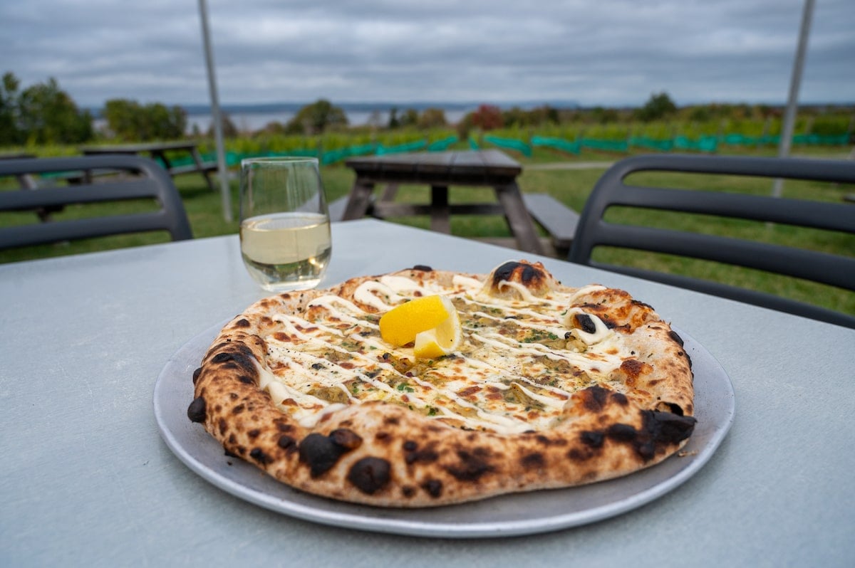 Wood-fired clam pizza topped with lemon, served alongside a glass of Tidal Bay 'L’Acadie' wine on an outdoor table at Lightfoot & Wolfville Vineyards, with vineyard views and picnic tables in the background.