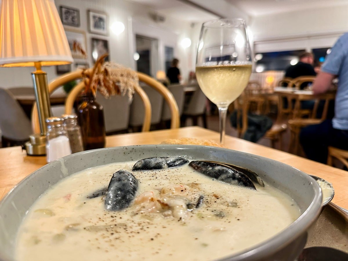 Close-up of a bowl of creamy seafood chowder with mussels, paired with a glass of local Nova Scotia white wine at Longfellow Restaurant, set on a cozy table with warm lighting and rustic decor in the background.