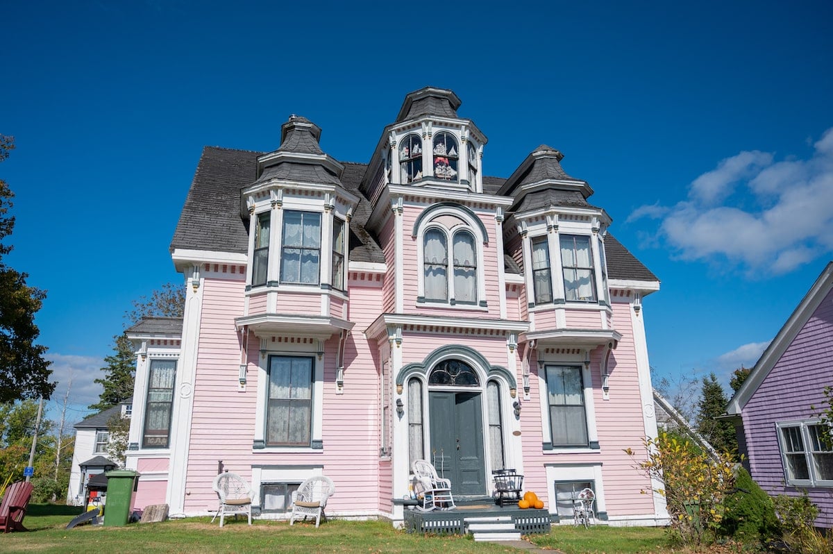 Front view of the historic 'Wedding Cake House' in Lunenburg in Nova Scotia, a pastel pink Victorian-style home with ornate white trim and bay windows, set against a clear blue sky.