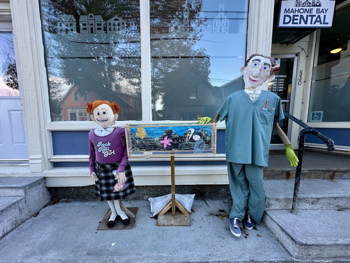 Two playful scarecrow figures on display outside a dental office at the Mahone Bay Scarecrow Festival, one dressed as a girl in a 'Rock n Roll Girl' shirt and the other in medical scrubs, standing in front of a fish tank display.