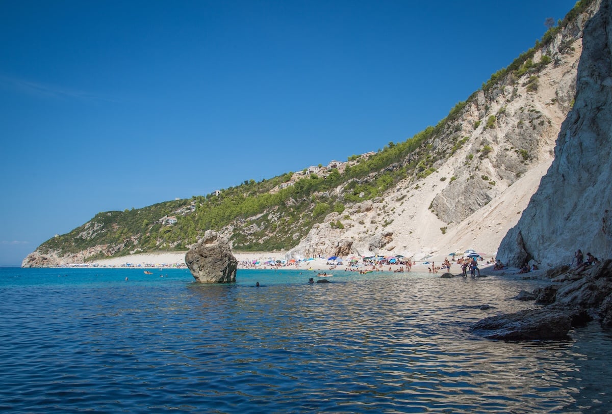 Milos Beach in Lefkada in the Greek islands, featuring clear blue waters with a large rock formation in the foreground, a sandy beach with sunbathers in the distance, and steep cliffs covered in greenery surrounding the area.