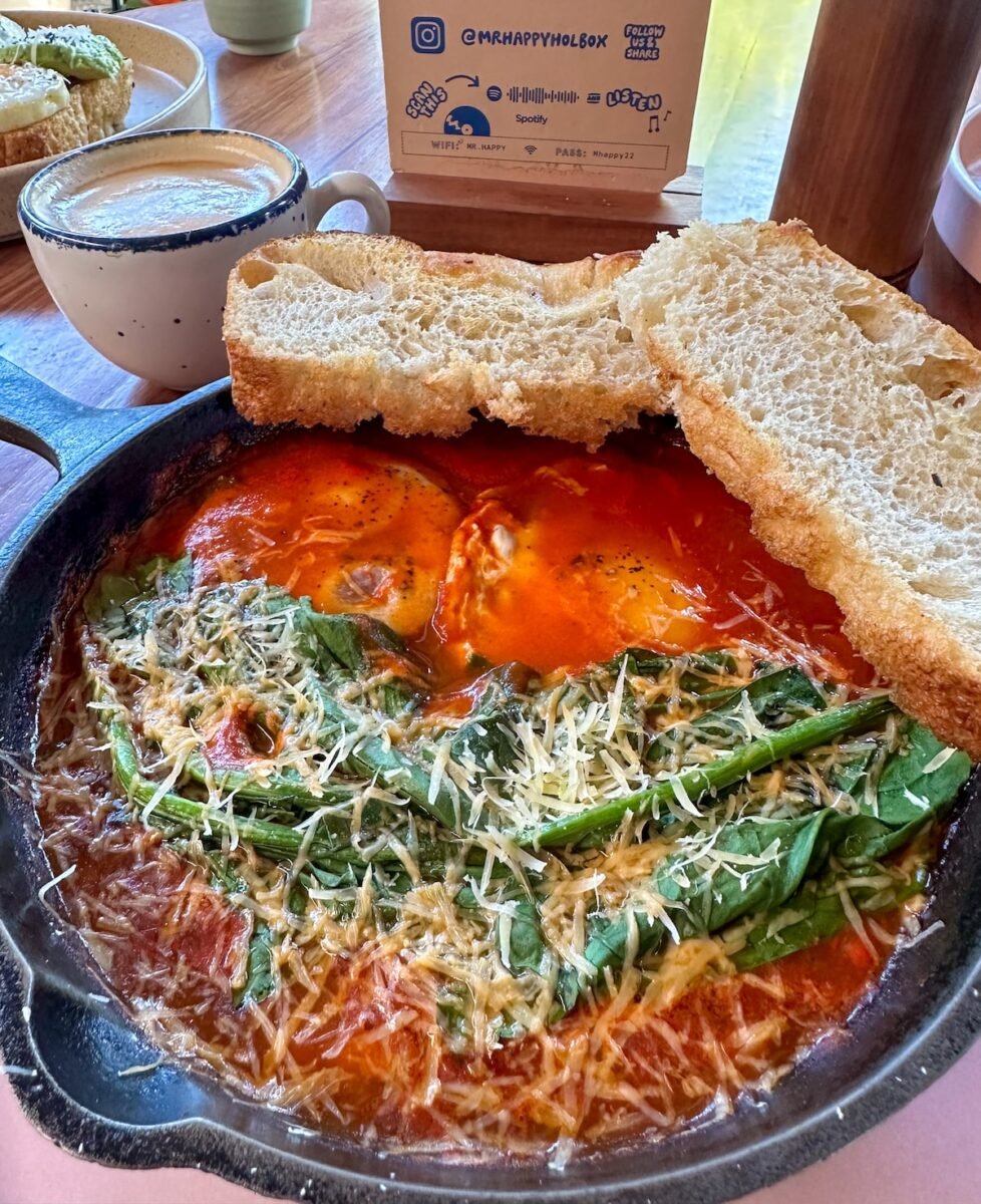 Close-up of baked eggs topped with fresh spinach and grated cheese in a rich tomato sauce, served in a cast-iron skillet with a slice of toasted bread on the side at Mr. Happy Holbox cafe.