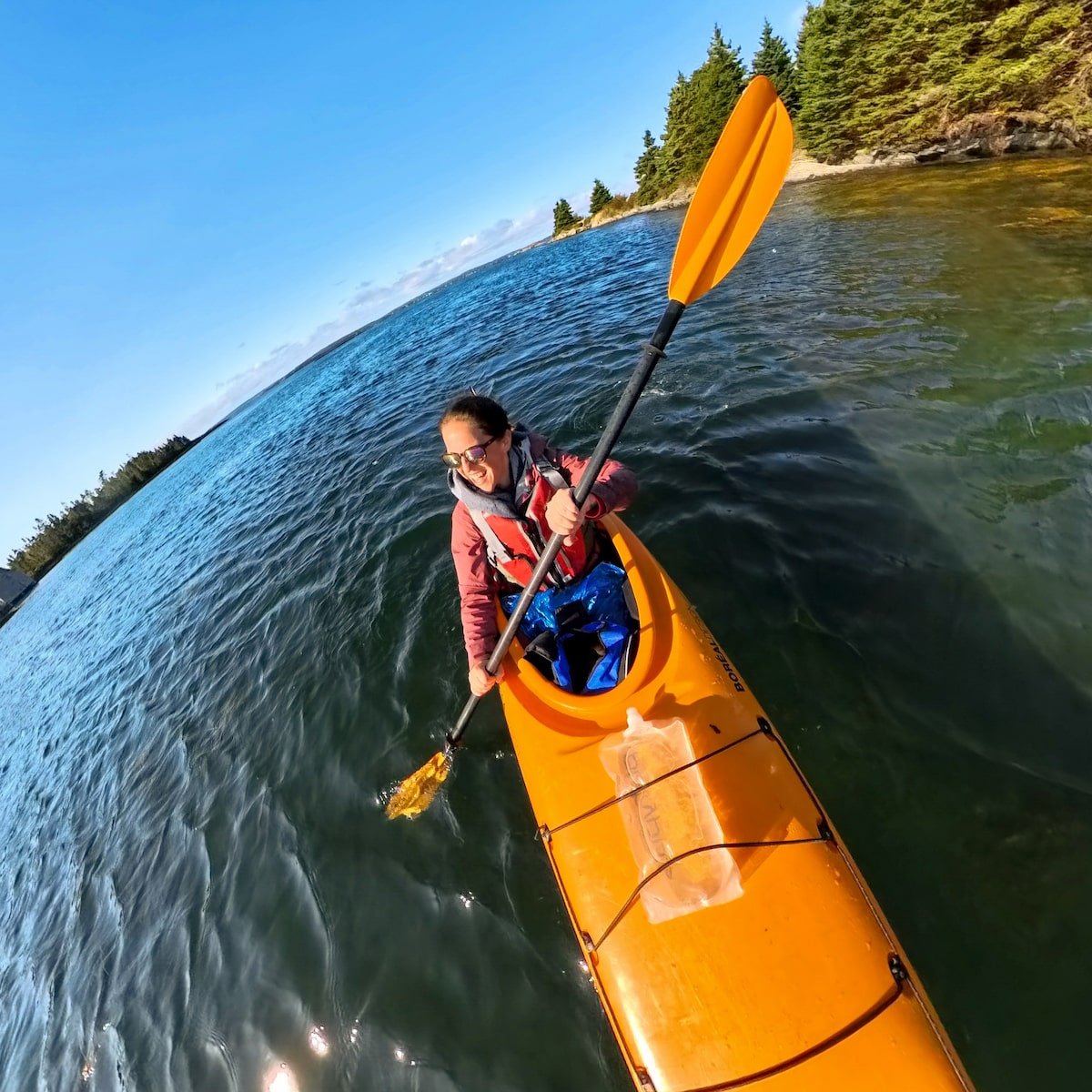 Smiling woman kayaking in the clear waters off Blue Rocks in Nova Scotia, paddling in an orange kayak with a backdrop of rocky shoreline and evergreen trees under a bright blue sky.