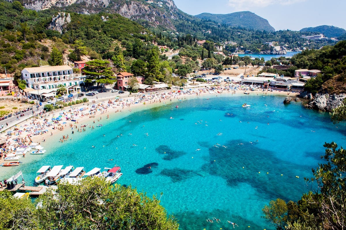 Palaiokastritsa Village in Corfu, Greece, with a view of the turquoise bay filled with swimmers, surrounded by a sandy beach lined with colorful umbrellas and buildings, set against a backdrop of lush green hills.