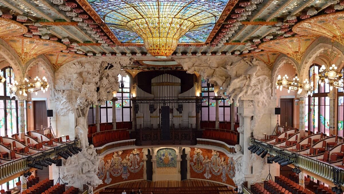 Interior of the Palace of Catalan Music in Barcelona, featuring a stunning stained-glass ceiling, intricate sculptures, and ornate decor surrounding the stage and seating area.