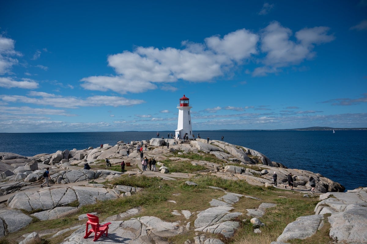 View of Peggy’s Cove Lighthouse in Nova Scotia perched on rugged granite rocks, surrounded by the Atlantic Ocean under a clear blue sky, with visitors exploring the iconic site.