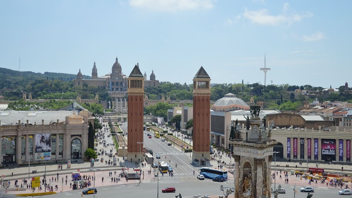 View of Barcelona's Plaça d'Espanya with the twin Venetian Towers in the foreground, leading up to the National Art Museum of Catalonia (MNAC) situated on Montjuïc hill, with bustling crowds and traffic adding vibrancy to the scene.