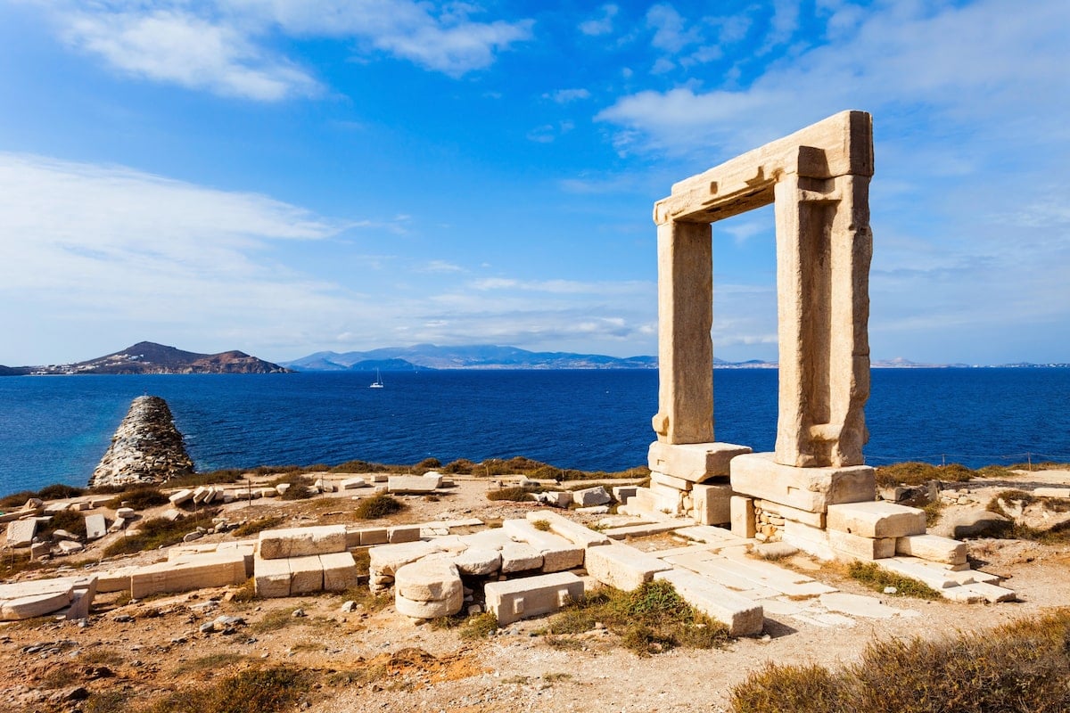 Portara Palatia on Naxos Island, an ancient marble gateway overlooking the blue Aegean Sea, with distant islands visible under a partly cloudy sky.