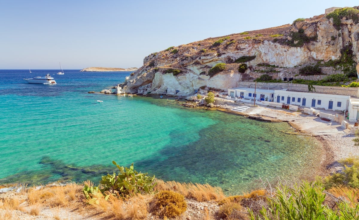 Rema Beach on Kimolos Island in Greece, showcasing crystal-clear turquoise waters, rugged cliffs, and a charming row of whitewashed buildings with blue doors. Boats rest in the calm bay, highlighting the secluded and serene atmosphere.