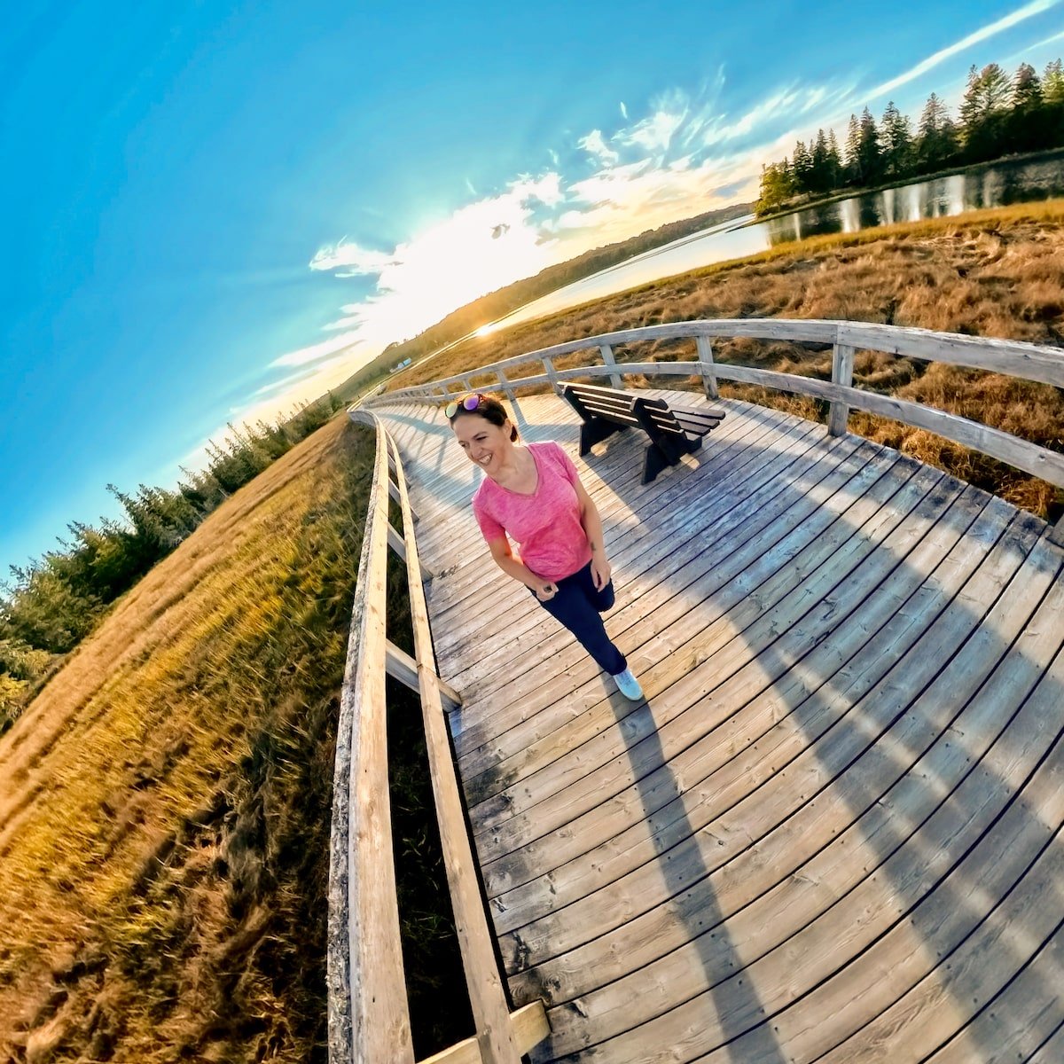 Smiling woman walking along the wooden Rissers Beach Boardwalk in Nova Scotia, with golden marshlands, a calm lake, and the sun setting behind the trees, casting long shadows on the boardwalk.