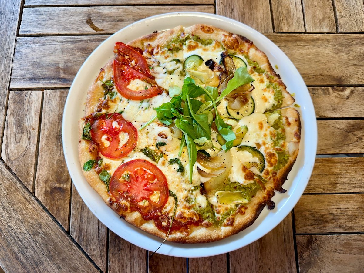 Pizza featuring two distinct halves: The Jo with garlic-parsley pea smash, roasted zucchini and onion, artichoke, red pepper flakes, and fresh arugula; and The Allyssa with marinara, mozzarella, freshly sliced tomato, arugula, and feta, served on a rustic wooden table.