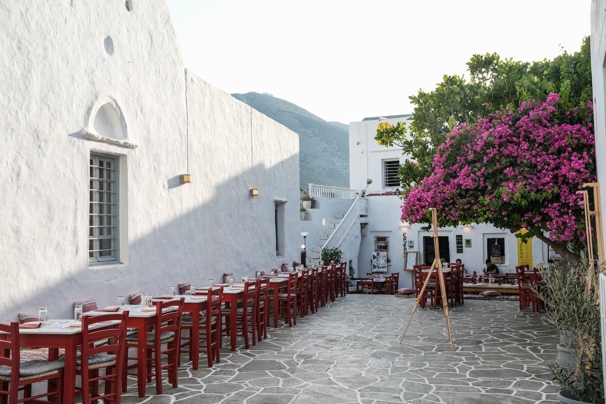 Outdoor restaurant in Sifnos with red wooden chairs, long communal tables, and vibrant pink bougainvillea covering part of the whitewashed building.