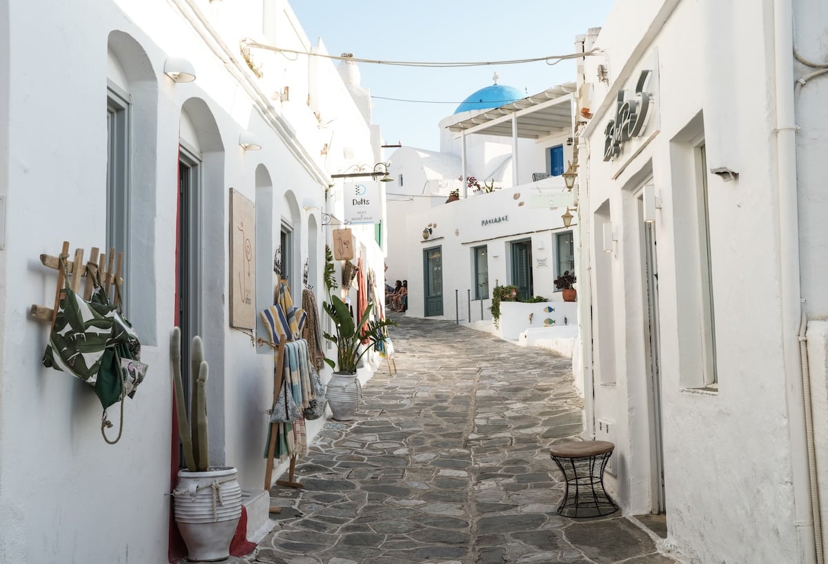 A charming alley in Apollonia, Sifnos, lined with whitewashed buildings, vibrant local shops, and potted plants. The iconic blue-domed church peeks over the rooftops, capturing the serene beauty and traditional architecture of the Greek islands.