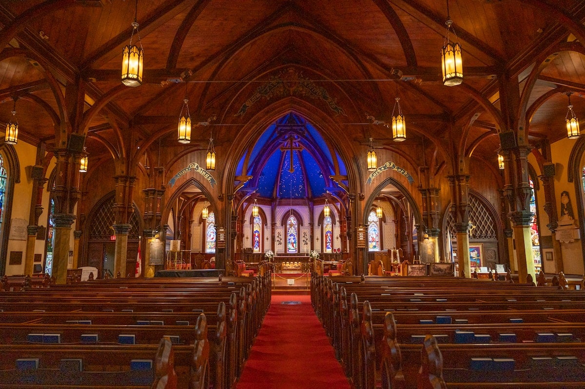 Interior of St. John’s Anglican Church in Lunenburg, featuring a high wooden ceiling with arched beams, dim lantern lighting, stained glass windows behind the altar, and blue accent lighting near the altar area, creating a warm, serene atmosphere.