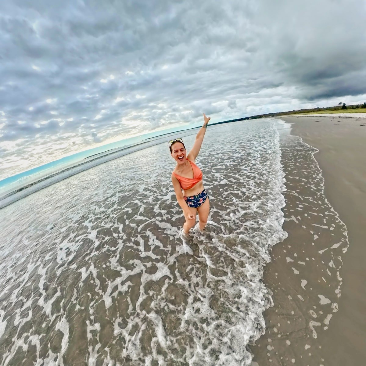 Excited woman standing in the shallow waves at Summerville Beach Provincial Park in Nova Scotia, wearing a bikini and smiling while raising one arm in the air, with the wide sandy beach and cloudy sky in the background.