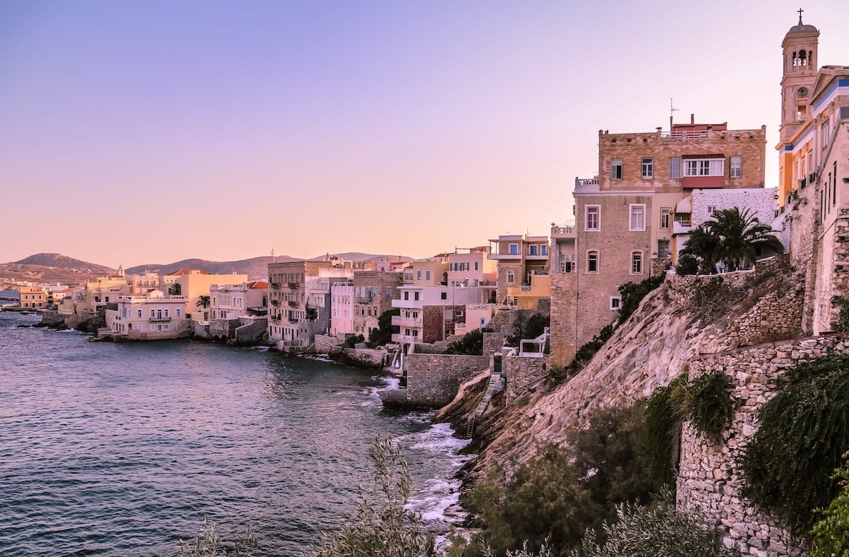 Sunset view of Syros, Greece, with pastel-colored buildings lining the rocky coastline and the sea reflecting the soft pink and purple hues of the sky.