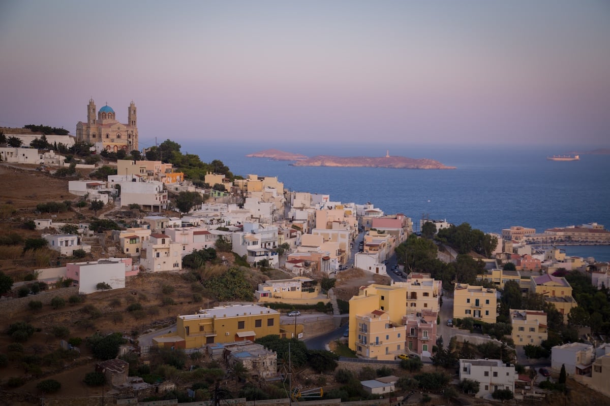 Solo traveler taking in a view from Ano Syros at sunset, showing pastel-colored buildings cascading down a hillside toward the Aegean Sea with a church perched on the left side and small islands in the distance.