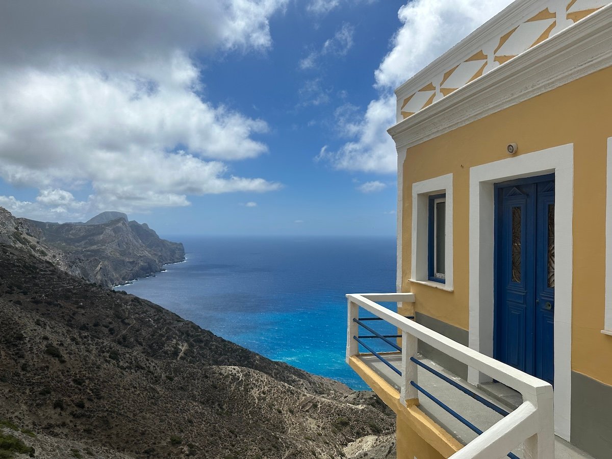 View of the rugged coastline of Karpathos in the Greek islands, with a traditional yellow and white house featuring vibrant blue doors and a balcony overlooking the expansive turquoise and deep blue waters of the Aegean Sea. The steep cliffs and rolling hills create a dramatic landscape under a partly cloudy sky, capturing the island's wild beauty.