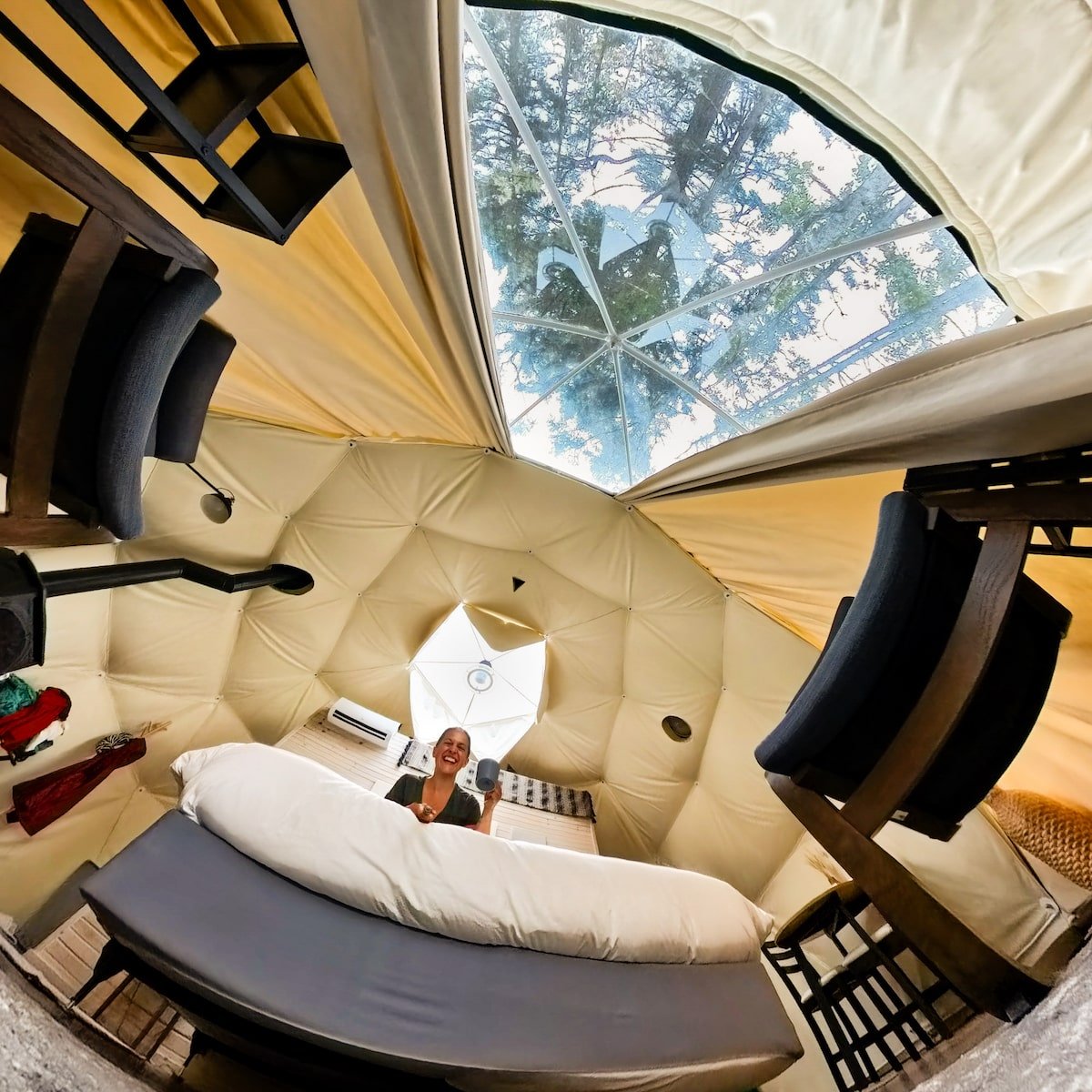 Cozy interior view of the Lakeside Glome at White Point Beach Resort, featuring a smiling woman holding a mug while sitting on the bed, with large windows in the dome ceiling offering a view of the trees and sky above.