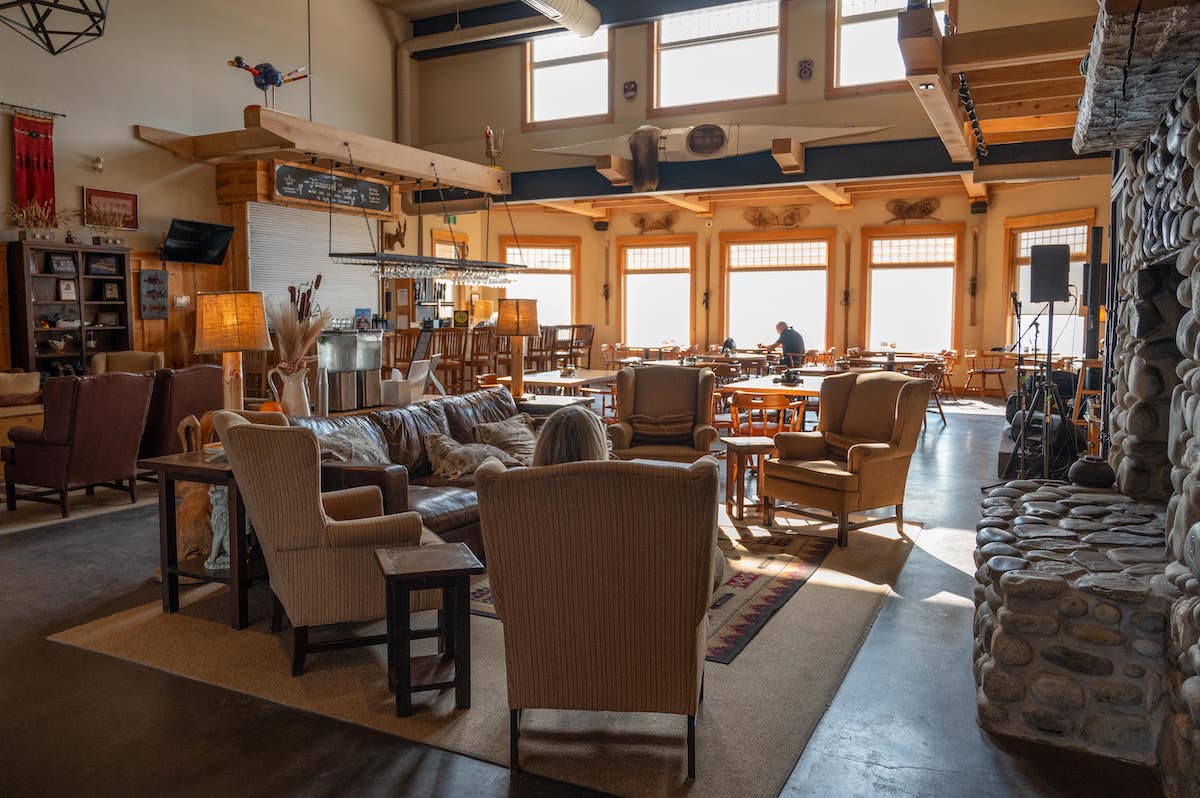 Cozy interior of the Founder’s Lounge at White Point Beach Resort in Nova Scotia, featuring comfortable armchairs, a leather couch, a large stone fireplace, and wooden tables with chairs, all illuminated by natural light from large windows.