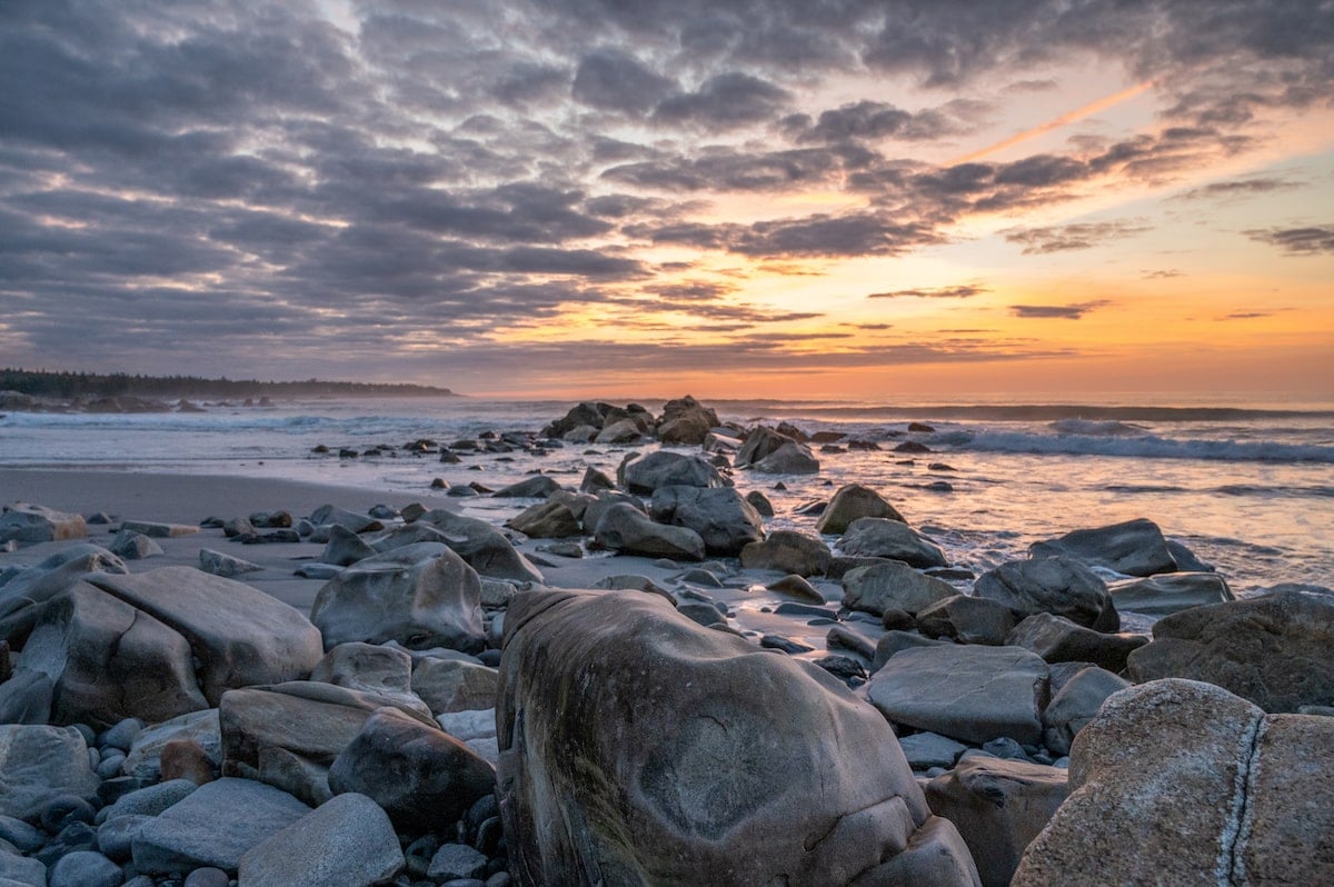 Stunning sunrise over White Point Beach Resort in Nova Scotia, with the sky painted in soft orange and purple hues, waves gently crashing on a rocky shoreline under a cloud-filled sky.