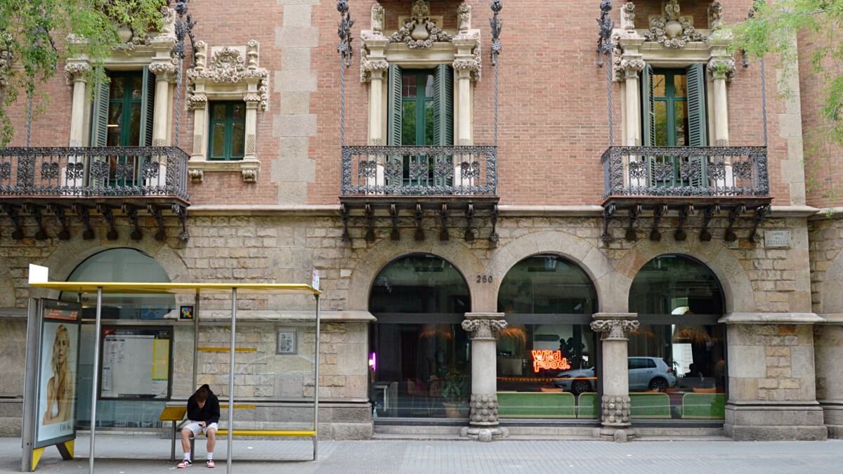 Barcelona street scene featuring a historic building with ornate balconies and arched windows. A lone person waits at a bus stop in front of the building, adding a touch of daily city life to the architectural beauty.