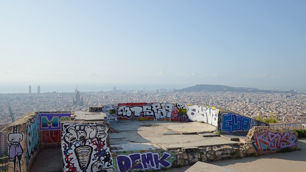 Panoramic view of Barcelona from the Bunkers del Carmel, with graffiti-covered walls in the foreground and a clear view of the city skyline and Mediterranean Sea stretching into the distance.