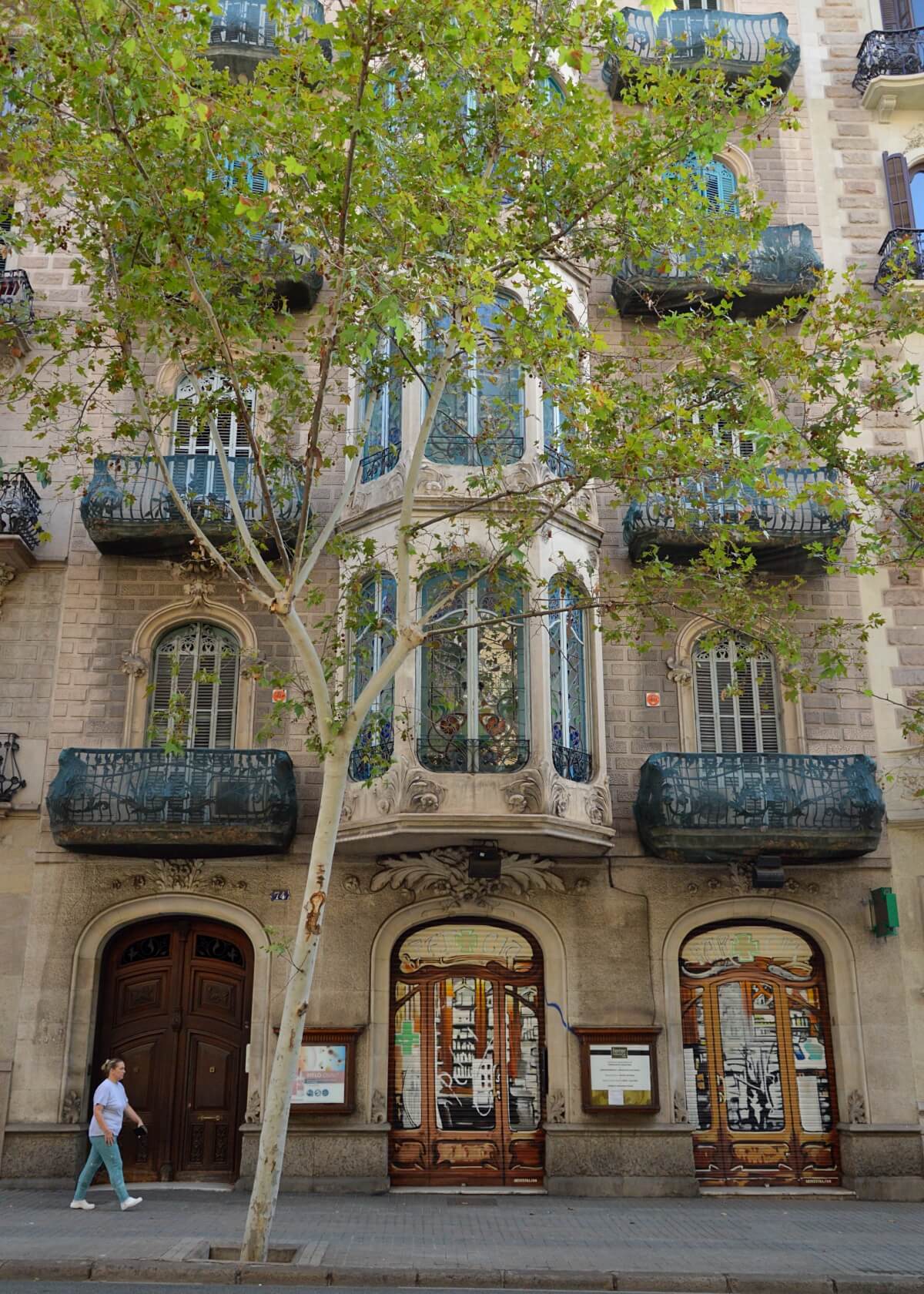 Street view in Barcelona's Eixample neighborhood, featuring ornate Art Nouveau architecture with decorative balconies, stained glass windows, and intricate door designs. A passerby adds a touch of life to the scene.