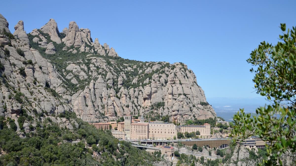 View of Montserrat mountain range with the Montserrat Monastery nestled at the base, surrounded by rugged limestone peaks and lush greenery under a clear blue sky.