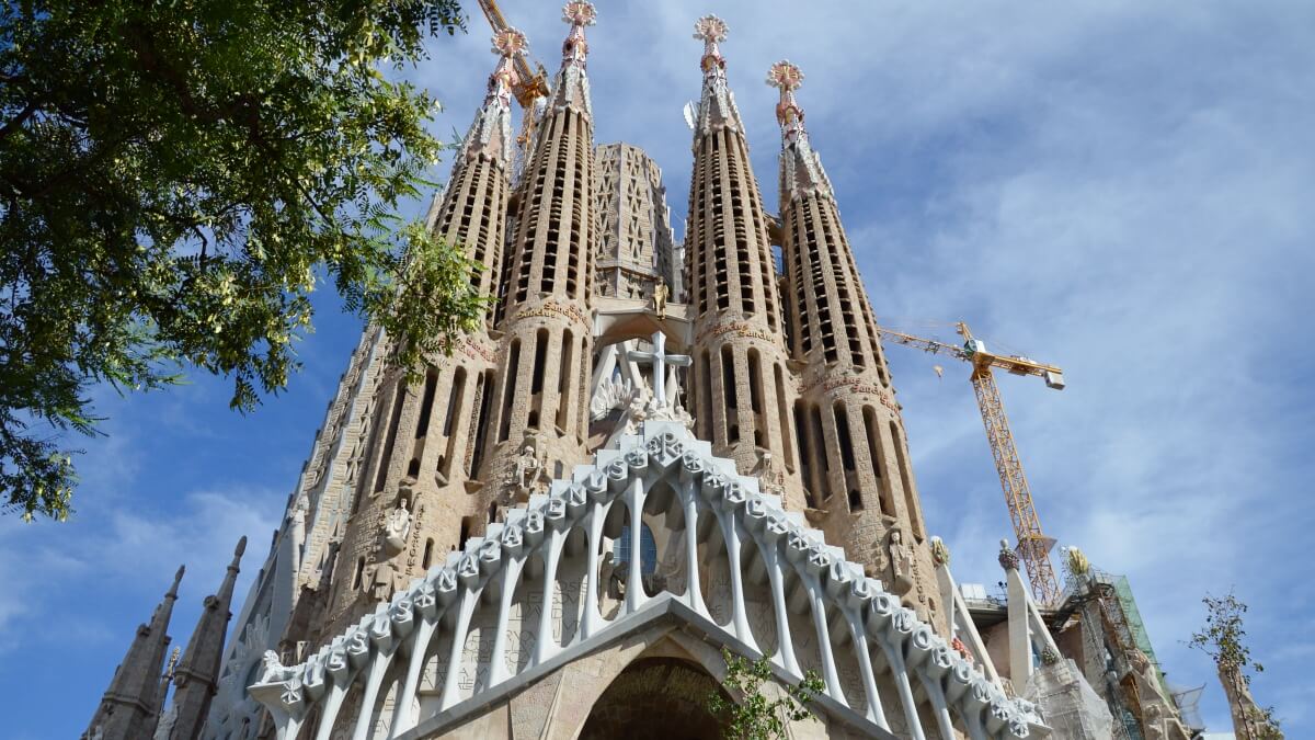 Close-up view of the Sagrada Família's intricate facade and soaring towers under a clear blue sky, with construction cranes visible as work on this iconic Barcelona landmark continues.