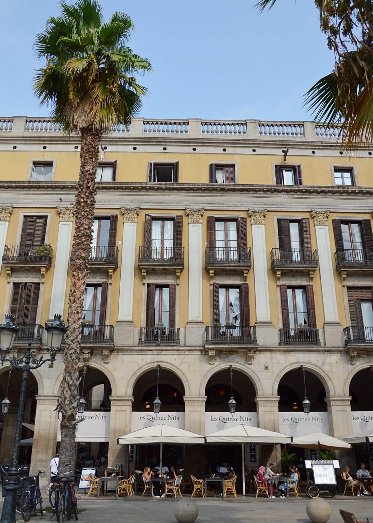 Outdoor seating at Les Quinze Nits, a popular tapas restaurant in Barcelona, with patrons dining under umbrellas in front of an elegant building with arched doorways and balconies.